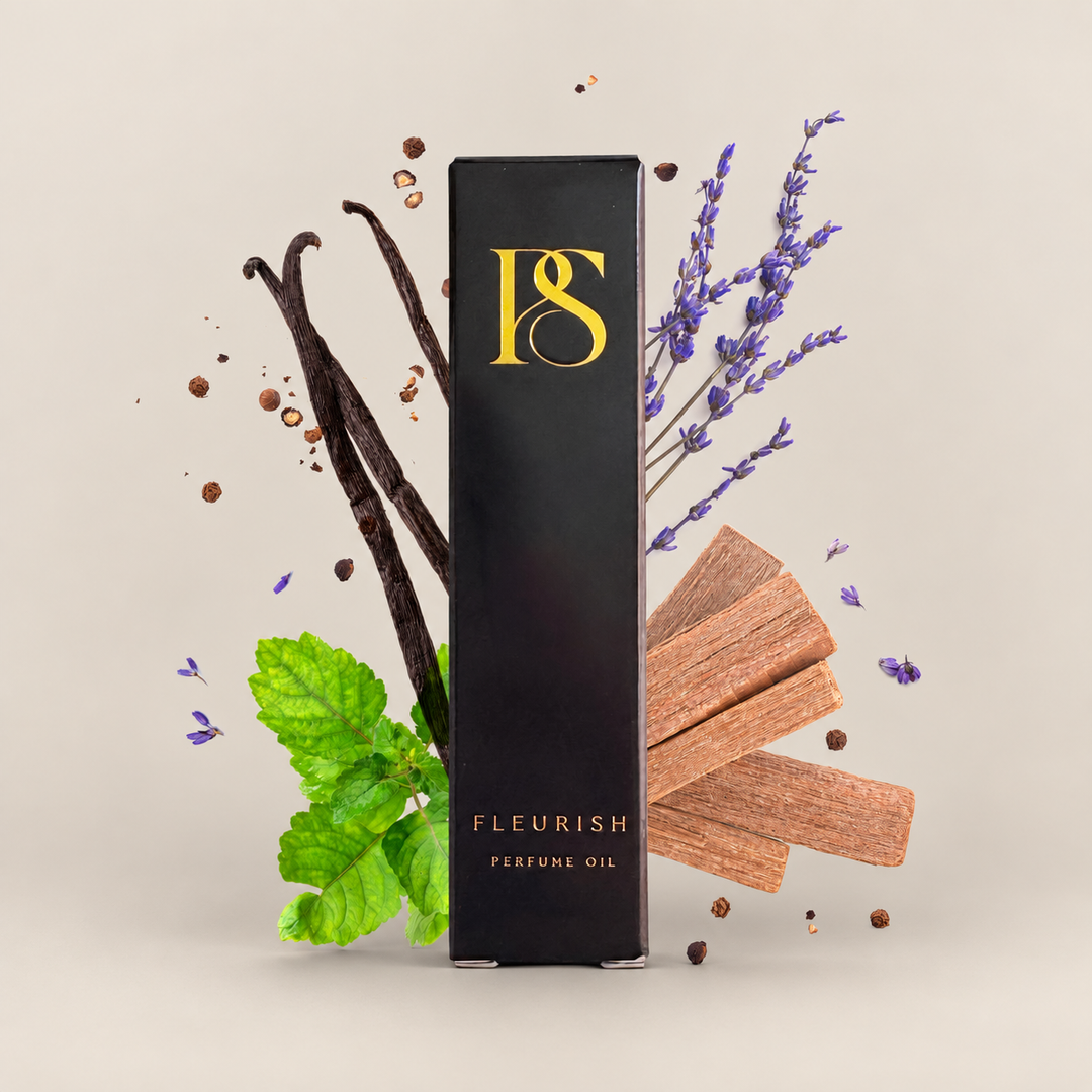A tall black rectangular box labeled 'FLEURISH PERFUME OIL' with a gold monogram logo stands centered on a light warm beige background, surrounded by fragrance ingredients including vanilla beans, lavender sprigs, mint leaves, and wood chips.