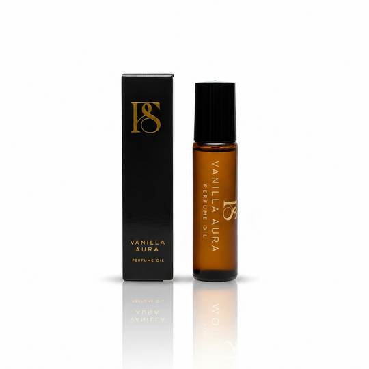 A bottle of Vanilla Aura perfume oil with a black cap stands next to its black packaging box, both featuring gold lettering and logo, on a clean white background with a soft reflection below.
